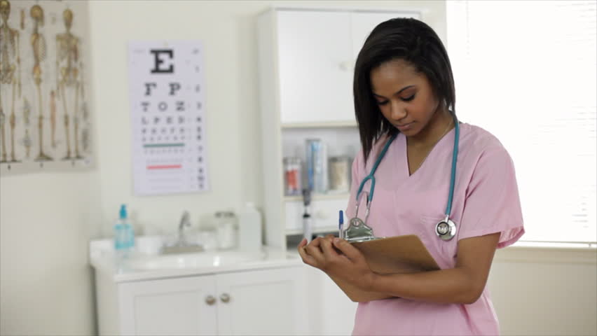A pretty young African American nursing student in a clinical setting stops writing on her clipboard and smiles at the camera.