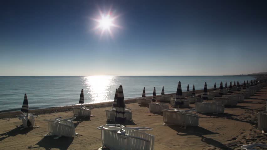 Many parasols and deckchairs standing on beach at sunny day, sun reflected in water