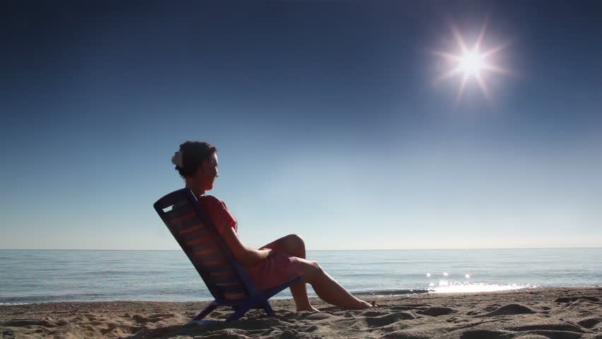 Woman sitting on deckchair and tans, on beach at sunny day, sideview