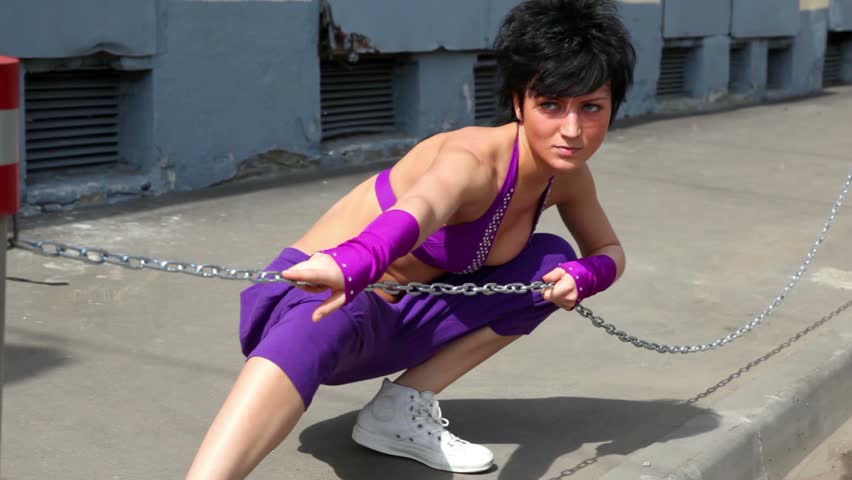 Woman in purple pants and a bra, make dance performance with chain, at pavement and industrial wall at Background