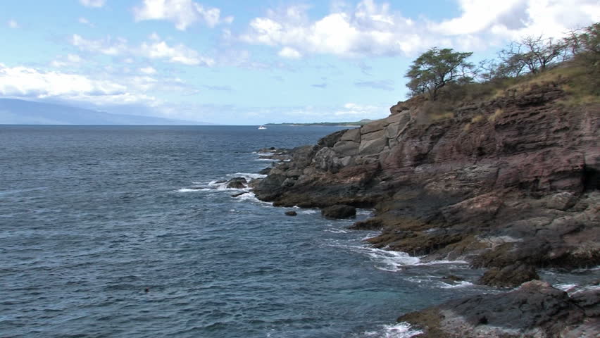 Rocky shoreline on Maui in Hawaii. A man swimming and spear fishing in the rough water. Cliff and trees overlooking water. Island of Lanai on horizon.