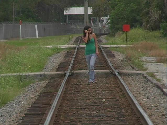 A beautiful young brunette in tattered jeans walks toward the camera on a lonely stretch of railroad track.  Long shot, full-length.