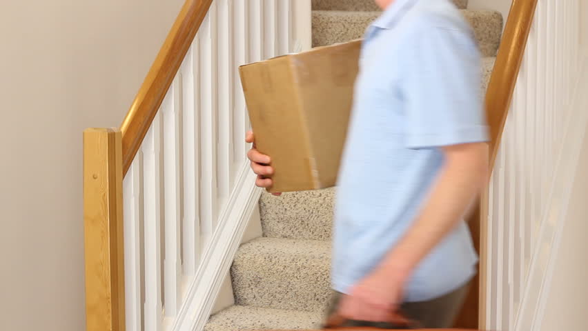 Mature male home from a business trip walking upstairs with luggage and a cardboard box.