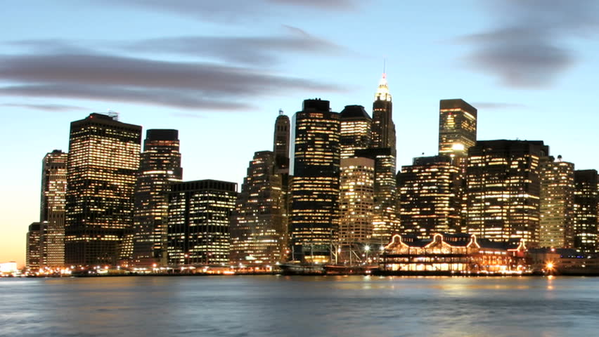 Slow pan of a time-lapsed shot of clouds in shades of pink and purple passing over the New York City skyline as golden-hour changes to night.