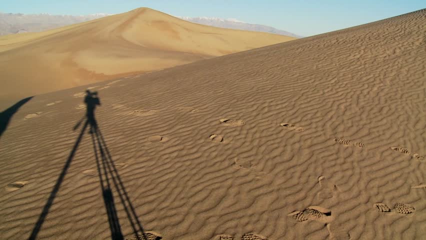 The shadow of a filmmaker with a tripod in shadow filming in Death Valley against the vast arid dunes.