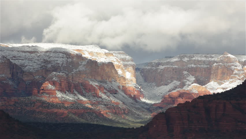 Sunlight shines down through heavy, early-morning clouds covering a mountainous canyon near Sedona, Arizona with a light dusting of snow.