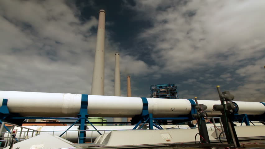 Wide dolly along white pipes and valves on the roof of a building with three massive smokestacks in the background at the IDE Ashkelon desalination plant in Israel.