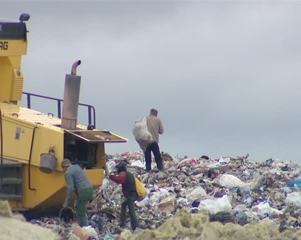 Poverty. Dump. Special machine pressing rubbish and homeless people looking for items and food. Environmental pollution. 