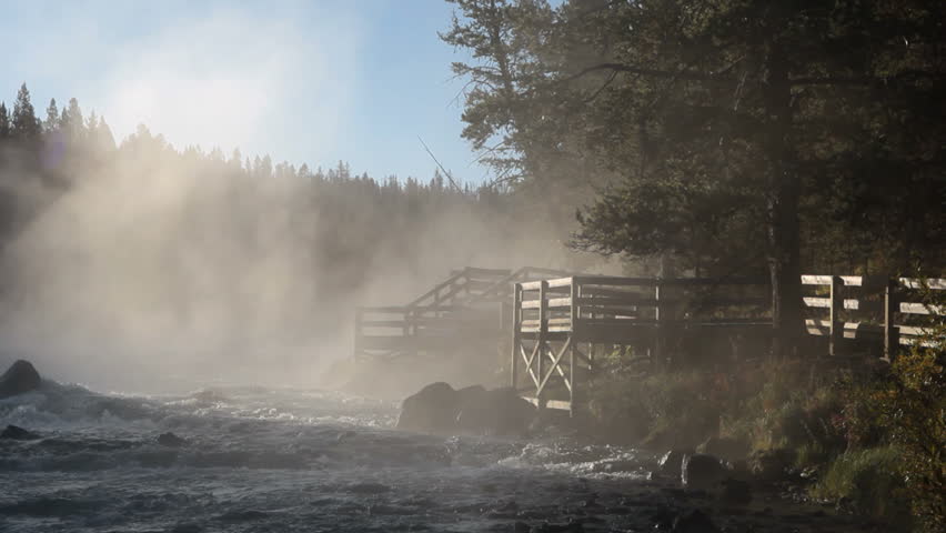 Yellowstone River with mist drifting near boardwalk overlook. Blue water, white mist and dark forest. Sunshine shining through with rays. Romantic and peaceful with a hint of mystery.