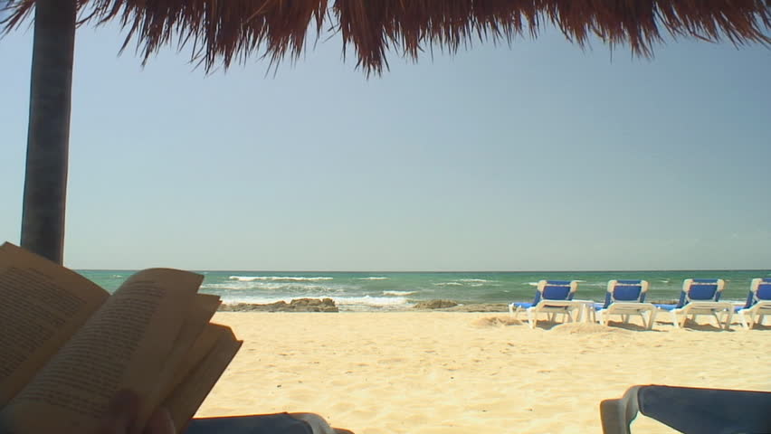 Book is read in shaded foreground as young couple strolls along sunny beach in background