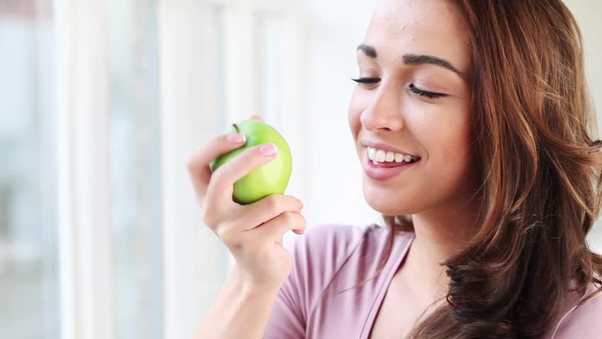 Healthy joyful  young woman eating green apple and smiling at home in front of window