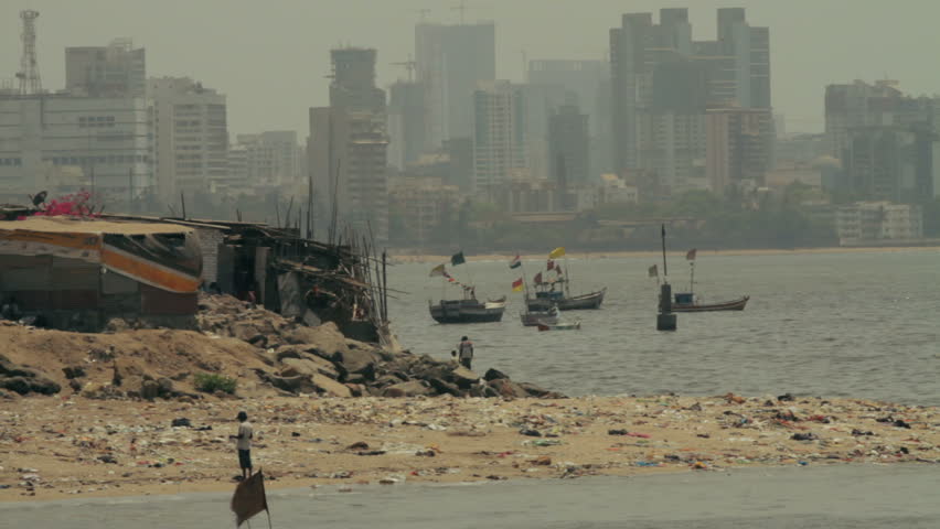 Locked off telephoto shot of child playing by the riverbank in Mumbai, India, with city skyline in the background.