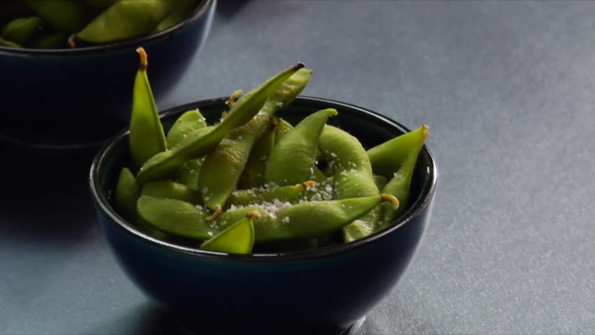 Close-up Pan up of man sprinkling salt on Edamame (soy beans) and then pouring steaming hot sake.