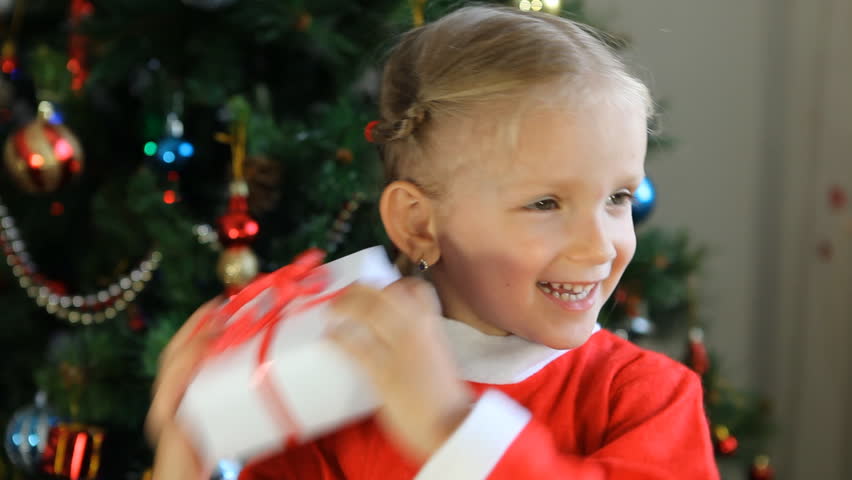 Child Shaking her Gift Near Christmas Tree, Happy Little Girl Dressed in Santa Claus Shaking her Gift Near Christmas Tree