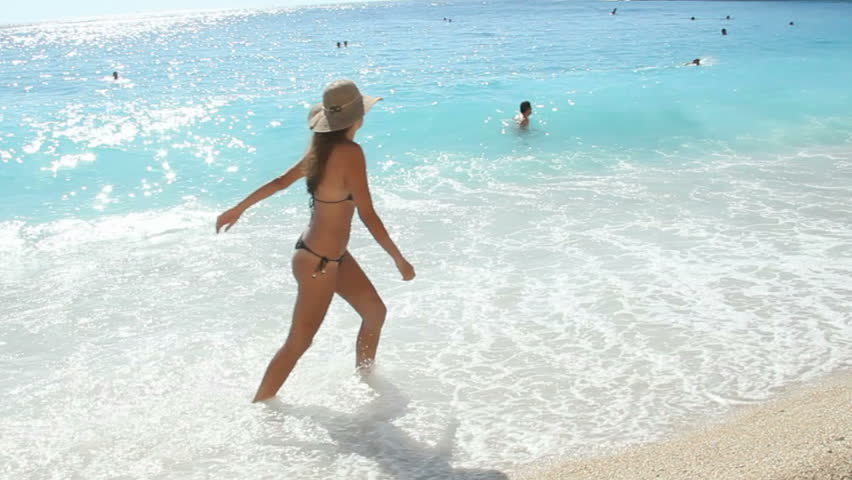 Tourist on a beach - Woman on vacation walking along the beach in shallow water, unrecognizeable people swimming in crystal clear turquoise blue water in the background