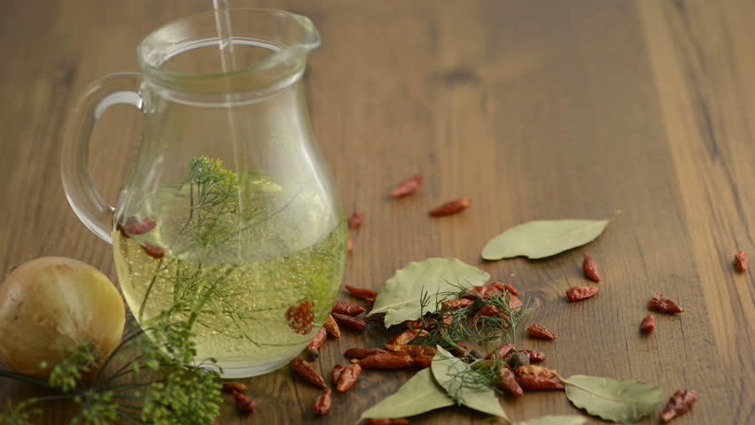 A carafe is filled with oil. Therein lie herbs such as dill and chili peppers. Outside are other spices such as bay leaves, onion, chili
