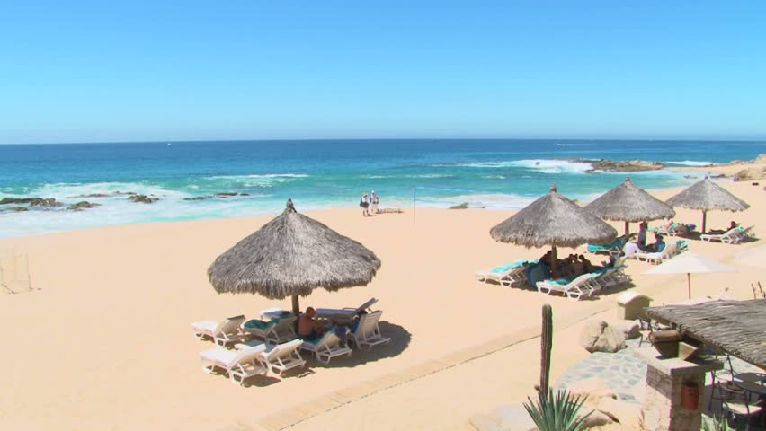People enjoy perfect blue sky day at the beach by tiki huts in Cabo San Lucas, Mexico by resort.