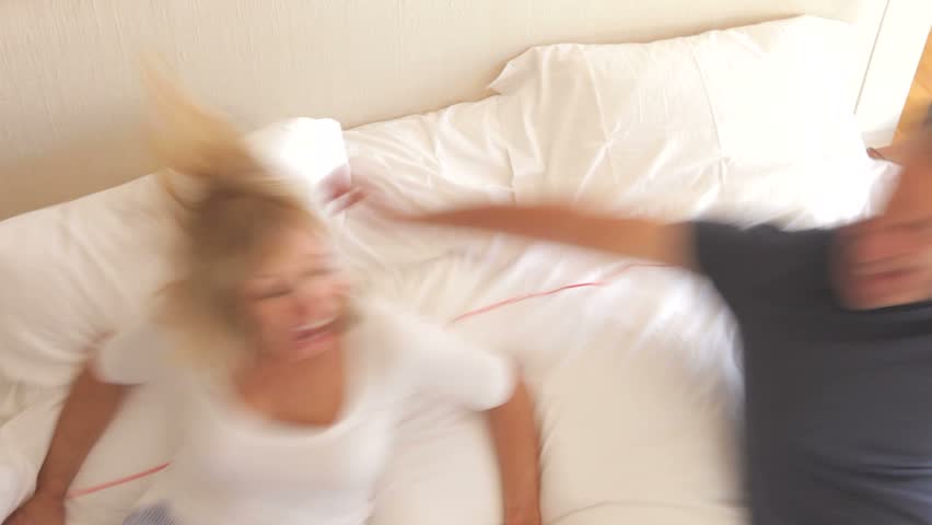 Senior husband and wife jump into frame and land on bed before looking at camera.