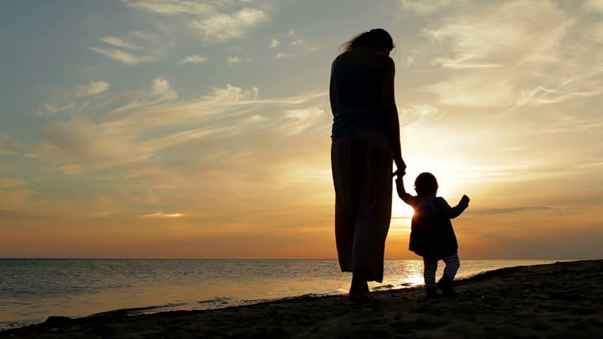 Mother with baby walking on sea coast. Silhouettes sunset.