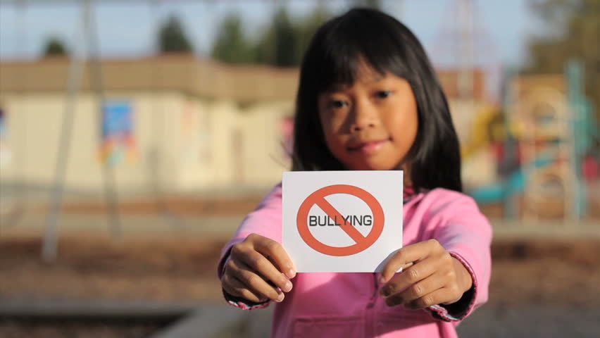 A cute Asian girl holds up a NO BULLYING sign in front of a school.