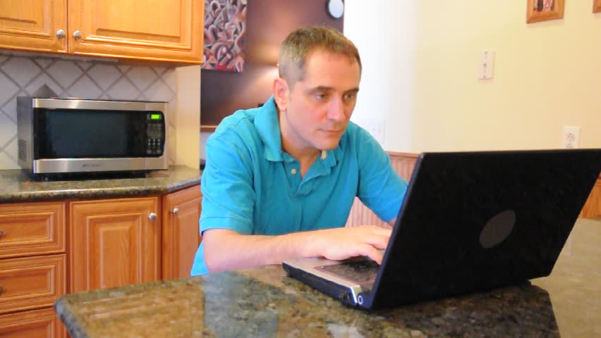 A casual man is sitting at his kitchen with a laptop and is frustrated at the computer because he needs technical help.