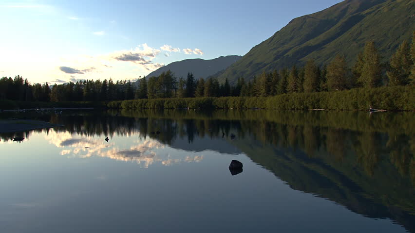 Late summer evening in Alaska - the sun beings to set in the west and the shoulder of a mountain and clouds reflect in the calm waters of a small lake in Portage Valley.