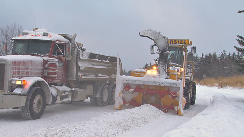 HOMER, AK - CIRCA 2011: Snow blower on an excavator paired with a dump truck to clean plow piles on snowy city street. Includes audio.