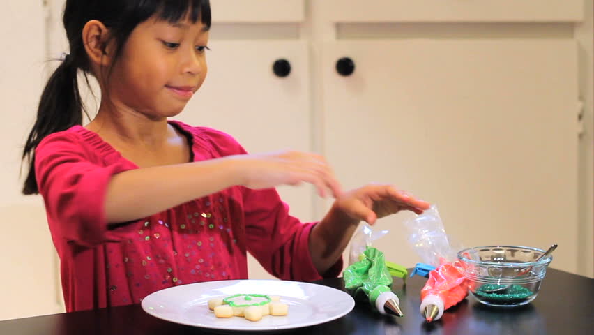 A cute little 6 year old Asian girl adds yummy red icing to her pretty Christmas cookie.