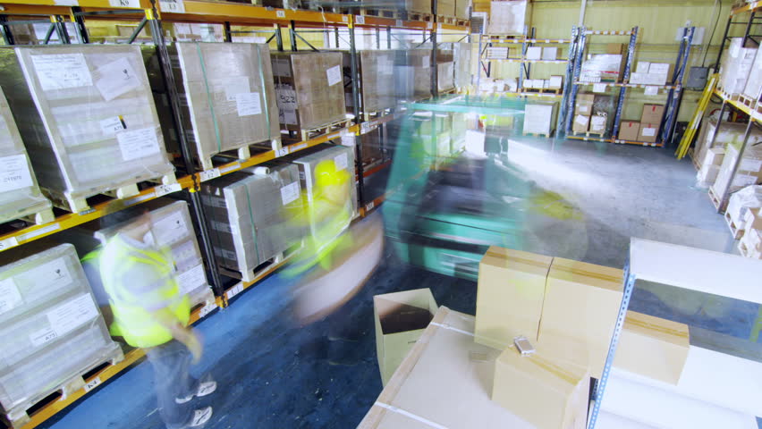 Time-lapse clip of workers in a warehouse in high visibility clothing and hard hats. They are moving boxes around and using a forklift truck to manoeuvre heavy stacks of goods around.