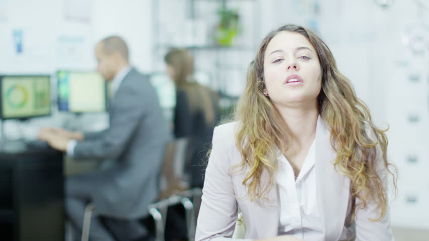 Portrait of a professional woman in business dress, she looks tired, stressed and irritable. 2 of her colleagues can be seen working in the background.