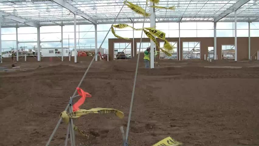 Clip of plastic yellow caution tape blowing in the wind inside large construction site with unidentifiable workers walking about.