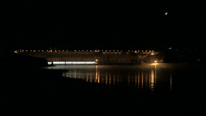 Dam of Krasnoyarsk Hydro-power Plant at night, long shot. Halogen illumination reflecting in water of Yenisei river, against black night sky with crescent. Divnogorsk, Russia. January 14, 2013.