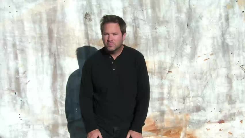 Man gestures with hand that he cannot hear and mouths words at same time, shot in bright sunlight with canvas backdrop.