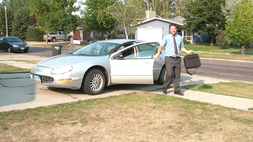 Businessman father pulls into driveway with car, and then comes out to wife, kids, and dog meeting him in yard, welcoming him home, in angled sun.