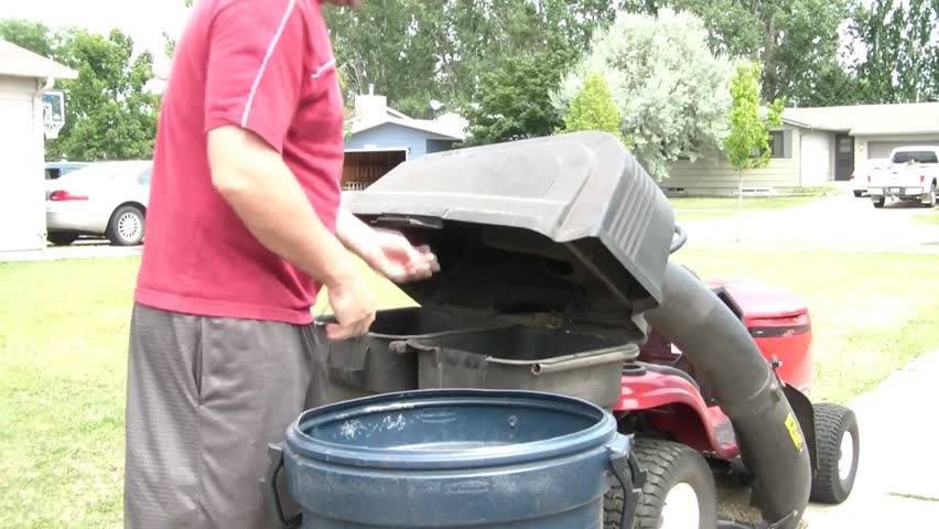 Guy stops red lawn tractor to unload grass from bagger into garbage can, in natural sunlight.