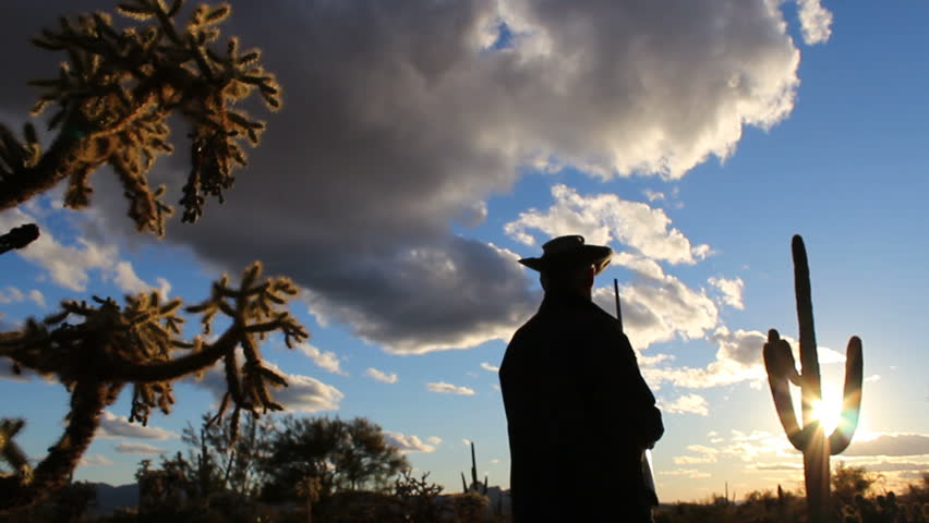 Arizona landowner holds rifle, walks, stealthily, searches for intruders in desert landscape at sunset. 1080p 