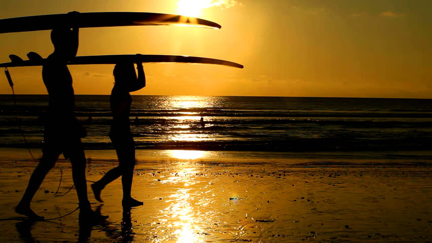 Surfer couple in silhouette walking with long surf boards at sunset on tropical beach