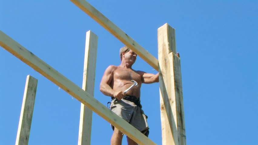Worker standing on crossbeam and hammering while building a barn. 