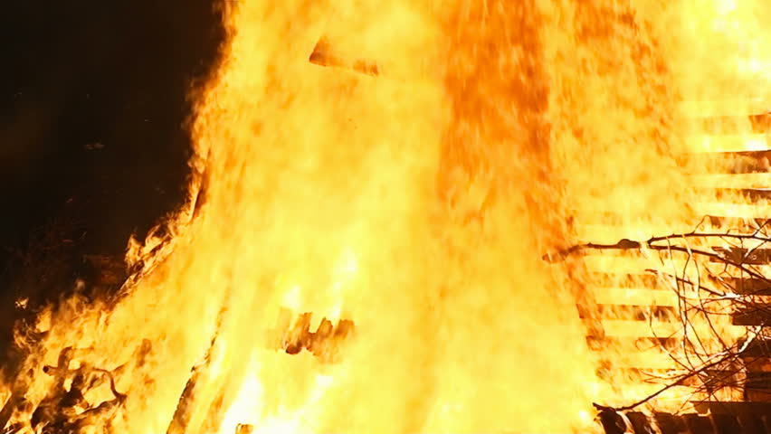 Fire from a big burning campfire, high flames. The flame pit is made of old wooden branches, the crackling trail is smooth over the dark black night. Slow camera movement towards the top of the pyre.