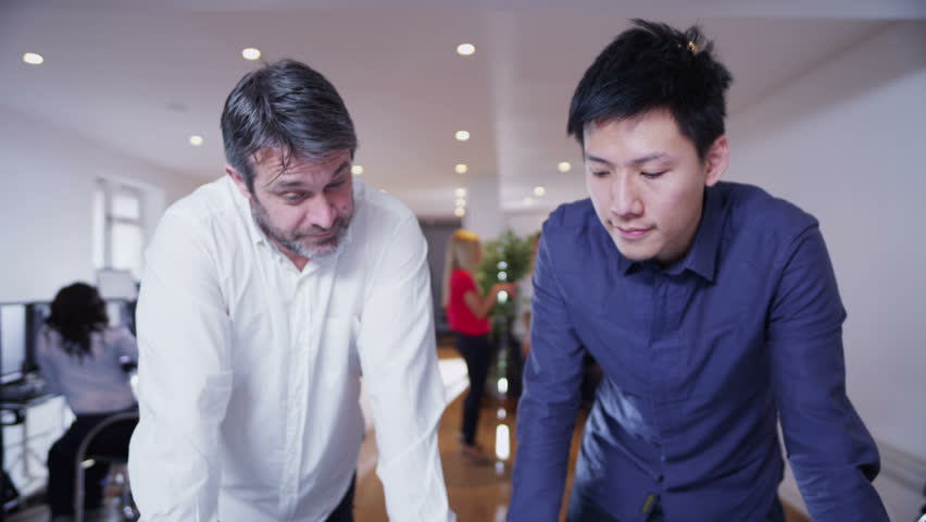 Two businessmen  of mixed ethnicity are working in a light and modern open plan office and looking at a map of the world. They are discussing possibilities for future business ventures. 