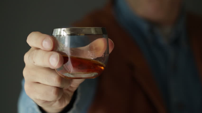 Whisky drinker wearing a denim shirt and leather vest presents a glass of whisky to the camera with his right hand. Recorded with macro lens and shallow depth of field.
