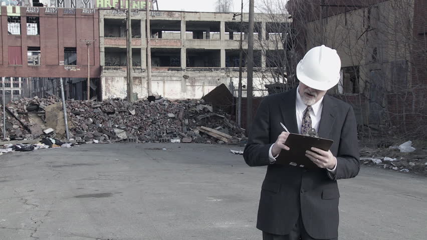 Developer (could be an architect, investor, contractor or other business person) walks into shot and looks around at the collapsed buildings around him, making notes. Medium long shot.