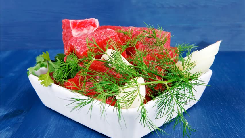 slices of raw fresh beef meat fillet in a white bowls with dill and green peppers serving over blue wooden table