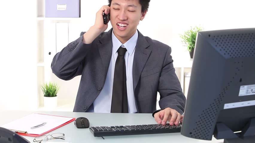 Young Asian businessman working on computer at his desk and picking up the phone, portrait