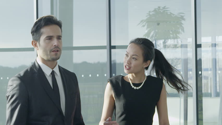An attractive mixed ethnicity businessman and businesswoman chat together as they walk around their light and modern office building. In slow motion.