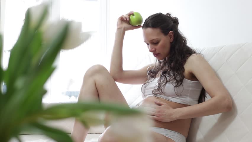Young pregnant woman sitting on sofa  eating green apple 