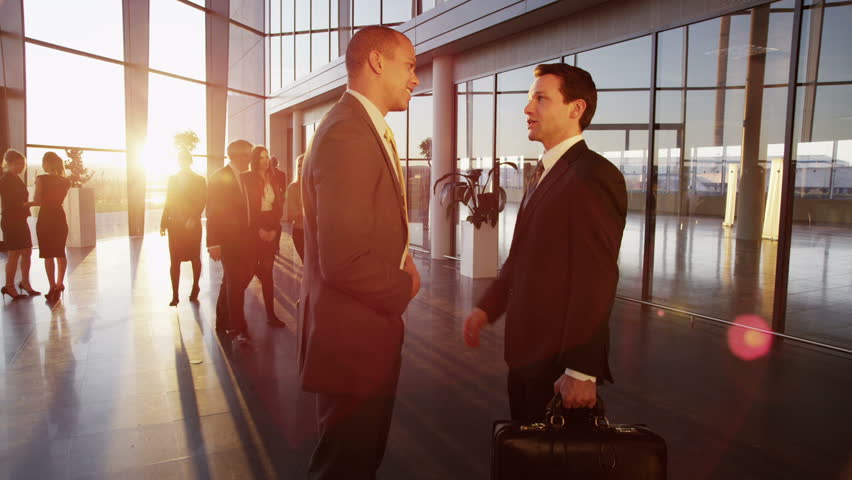 Two young businessmen who are old acquaintances, meet and shake hands in a busy modern office building at sunset. 