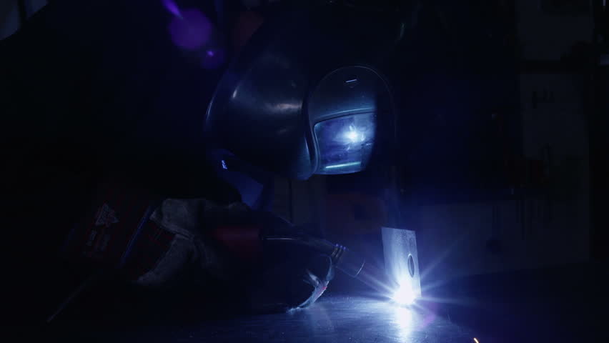 A welder is working in a dark construction or engineering plant. The flare of the flame is reflected in the visor of his mask.