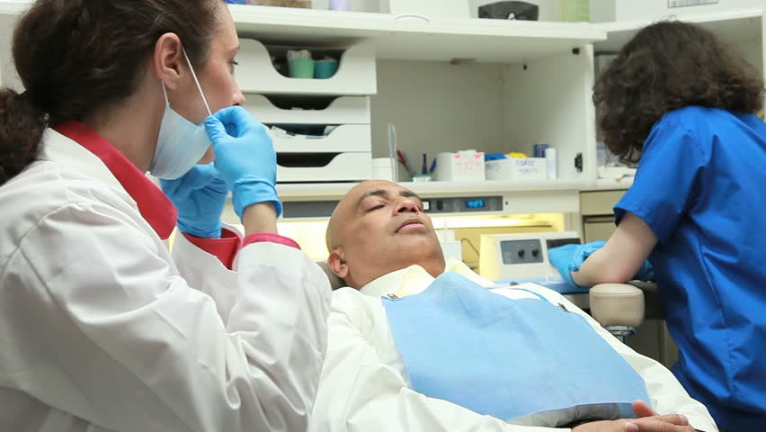 A female dentist receives a dental implement from her young intern assistant prior to commencing to examine the male patient in the exam chair.