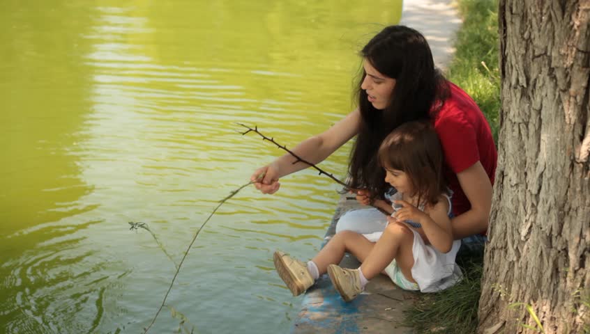 Brunette young adult mother teaches little baby daughter fishing.
